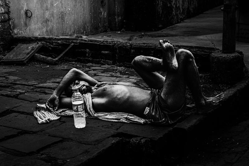 The monochrome street holds a reclining figure beside a plastic bottle, his stillness set against the paving and passing shadows.