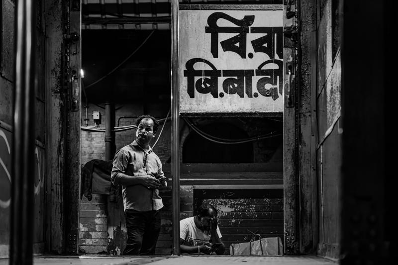 A woman stands below Bengali lettering at the entrance, her sari and pale face framed by the shop’s deep shadow.
