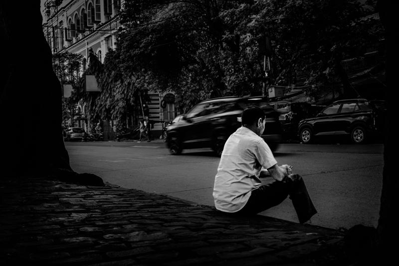 A man sits beneath the trees beside passing traffic, his turned back and still posture set against the blurred street.