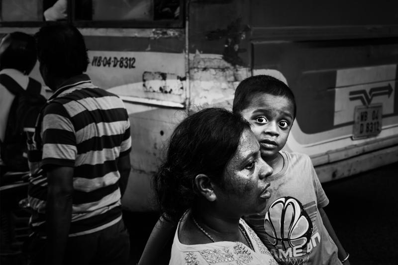 A child peers over a woman’s shoulder beside the truck, the bright face drawing attention through the cramped, shadowed street.