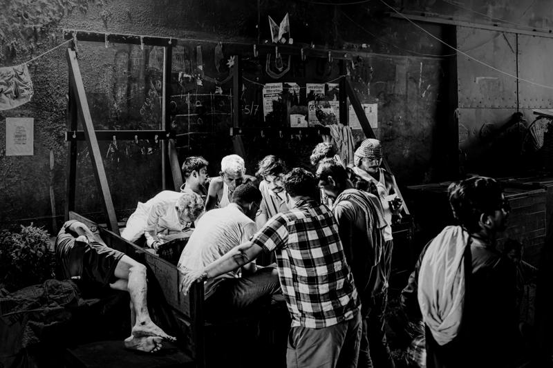Men lean over a low counter to inspect goods, checked shirts and bent shoulders forming a circle in the lane.