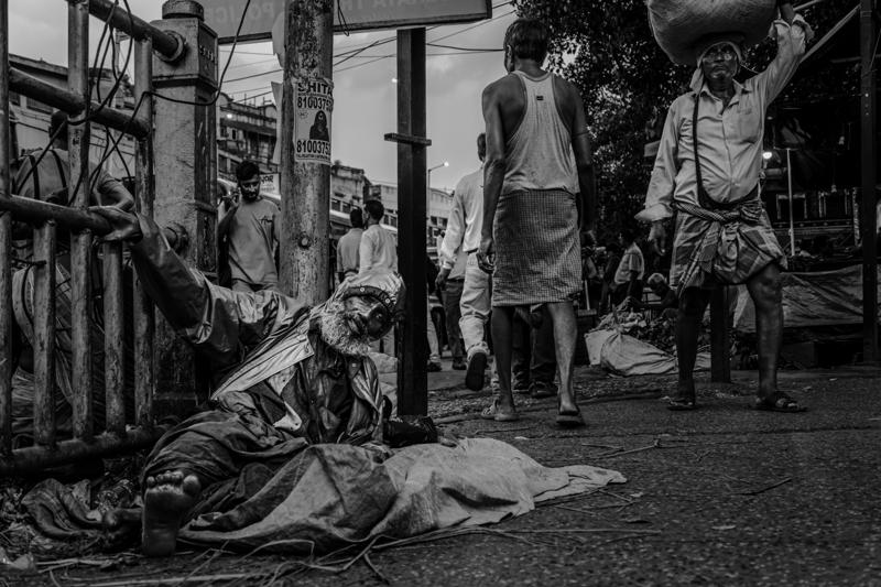 A man sits on the pavement beside his load while shoppers walk past and people stand near the market entrance.