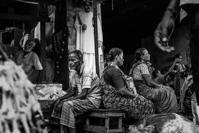 Several men sit shoulder to shoulder beside their baskets, their still figures framed by the dark market entrance behind them.