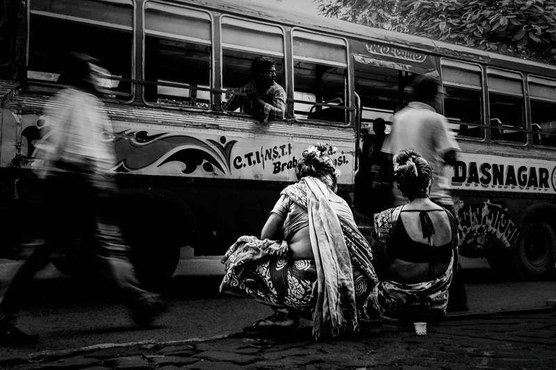 Women sit beside the road with bundles, while a bus fills the background and shoppers pass through the monochrome street.