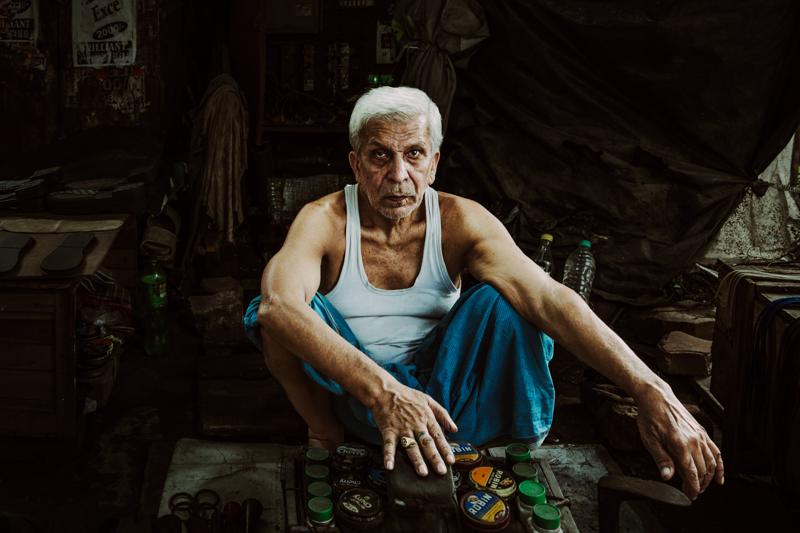 An older man sits in the shadowed stall, his turquoise trousers against the quiet clutter and black doorway around him.