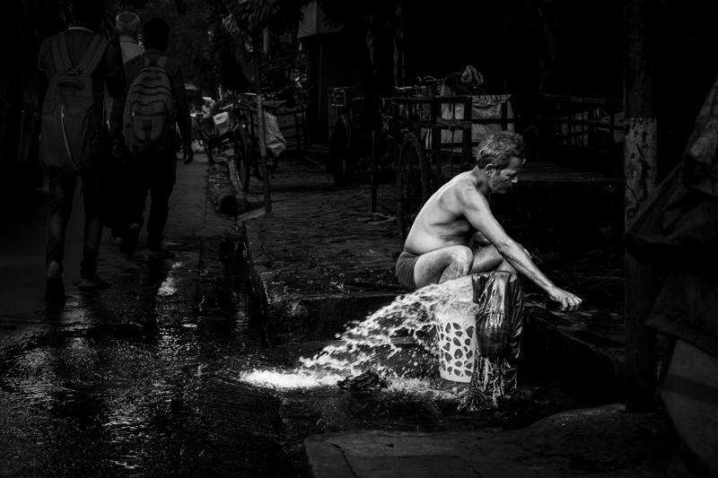 A worker bends over a basin in the wet street, beside buckets, scattered produce and dim market stalls behind him.