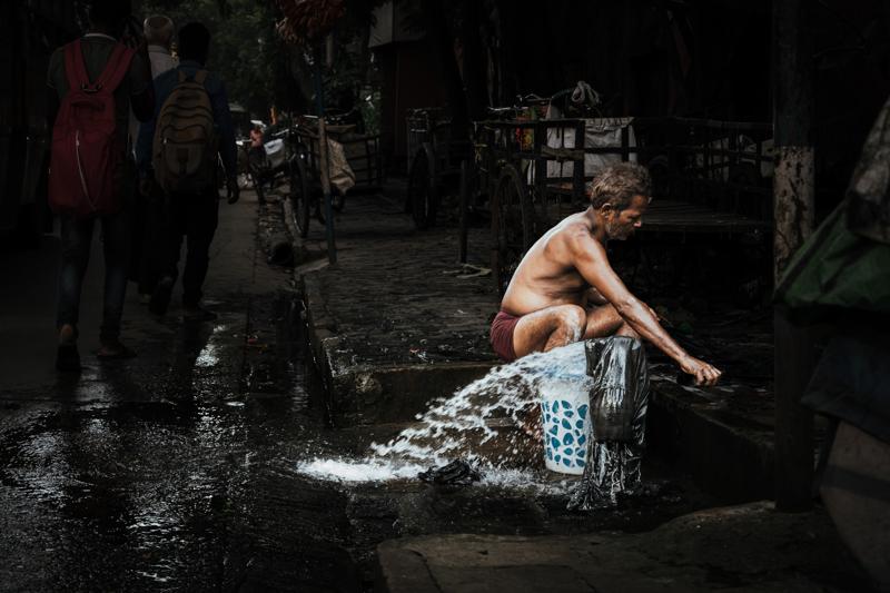 A man crouches beside a puddle to wash a patterned cloth, his hands and the rippling water against the lane.