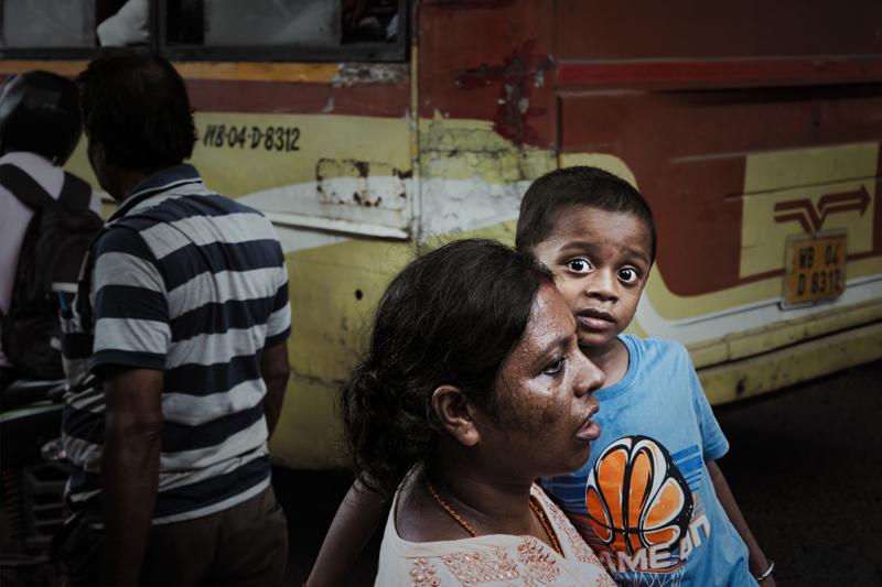 A mother holds a child who looks toward the camera, while pedestrians pass the yellow vehicle along the market street.