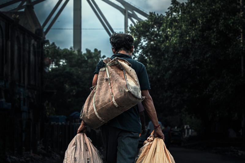 A man walks away with woven bags in both hands, moving past the bus and storefronts toward the market crowd.