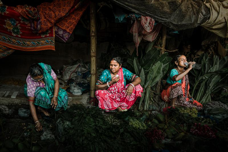 Several women sit together beneath dark cloth, their bright saris and small baskets filling the market stall with colour throughout.