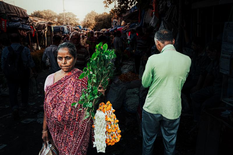 A woman in a pink sari holds a bundle of blossoms between market shoppers, the stems against the packed street.