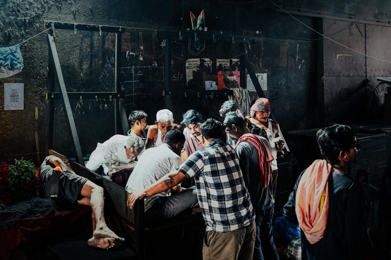 Several men lean over a market table, sorting items while their checked shirts and close shoulders form a busy cluster.