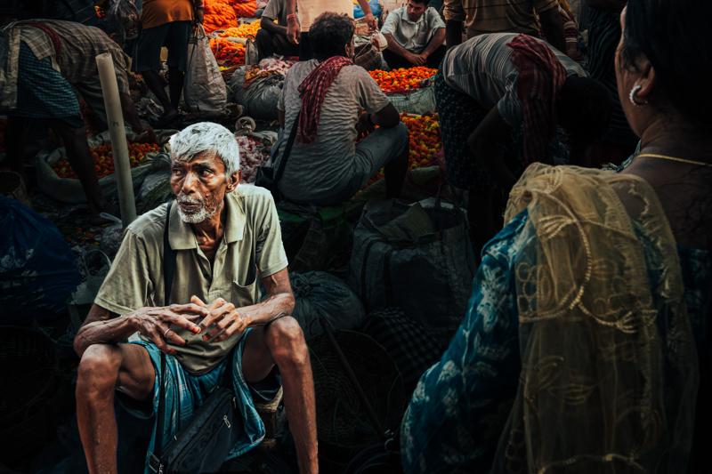 A grey-haired vendor sits with hands clasped while shoppers press around him, his pale shirt distinct amid the market’s colours.