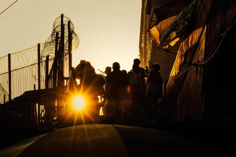 The low sun shines directly through the steelwork, turning passersby into silhouettes along the narrow riverside path, nearby, at dusk.