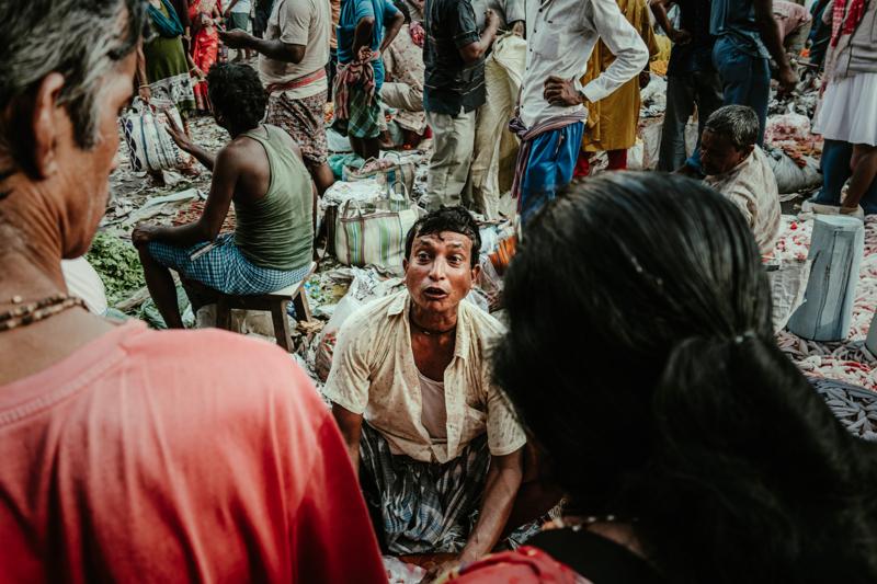 A man leans across the counter as customers in around him, their faces and hands layering the crowded market frame.
