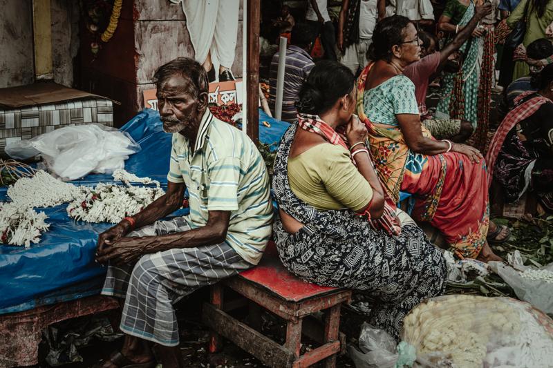 Several women sit among baskets and goods while one seller leans toward a customer, their saris in the market light.