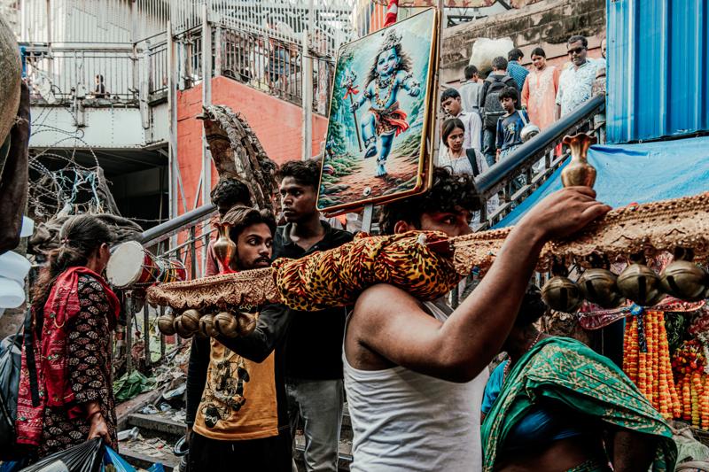 Two shopkeepers shift bundles beneath hanging stock, their bent postures and close quarters framed by the packed storefront.