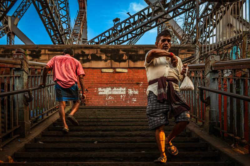 Two people cross the bridge approach between rusted beams, their bright clothing and measured steps set against the dense steelwork.