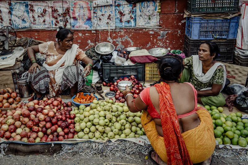 Vendors and customers cluster around produce baskets beneath a shop sign, while a shopper in a red shawl crosses nearby.