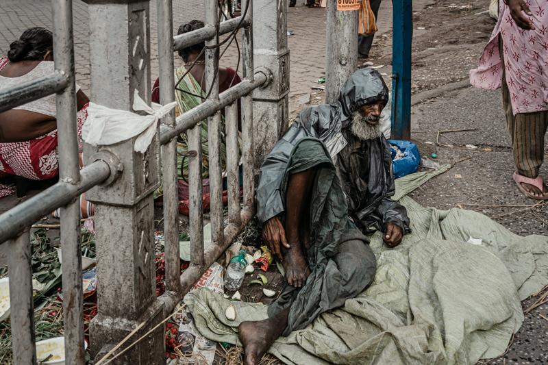 A man sits against a bamboo fence beside bundled sacks, while another person stands nearby along the uneven market street.