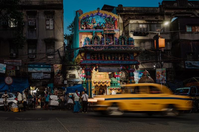 A yellow taxi blurs past pedestrians and shopfronts with painted signs and hanging goods lining the market lane at night.