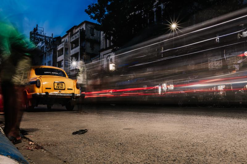 Red and white light trails streak past a yellow taxi on the night road, while dark storefronts frame the traffic.