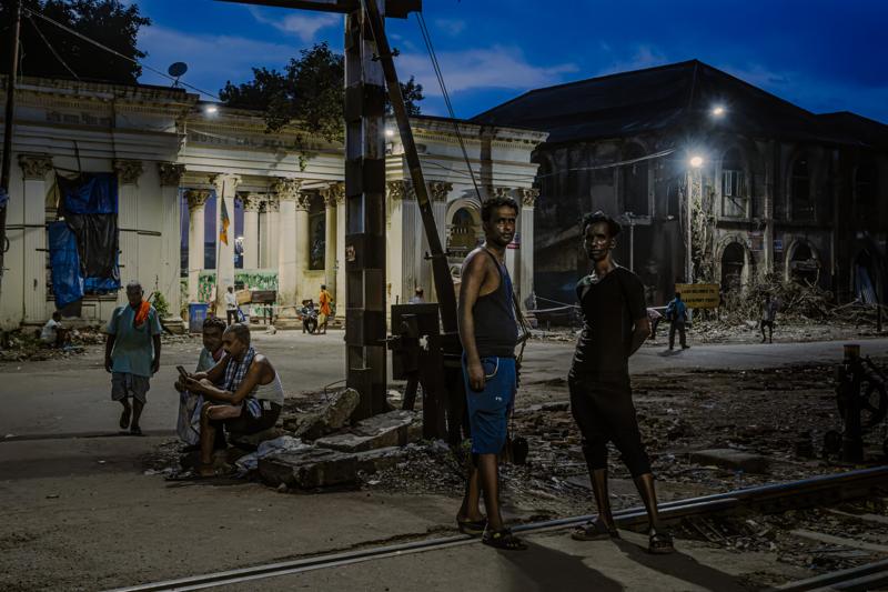 Pedestrians cross the lane past a lit market entrance, while a woman carries a basket through the busy street crowd.