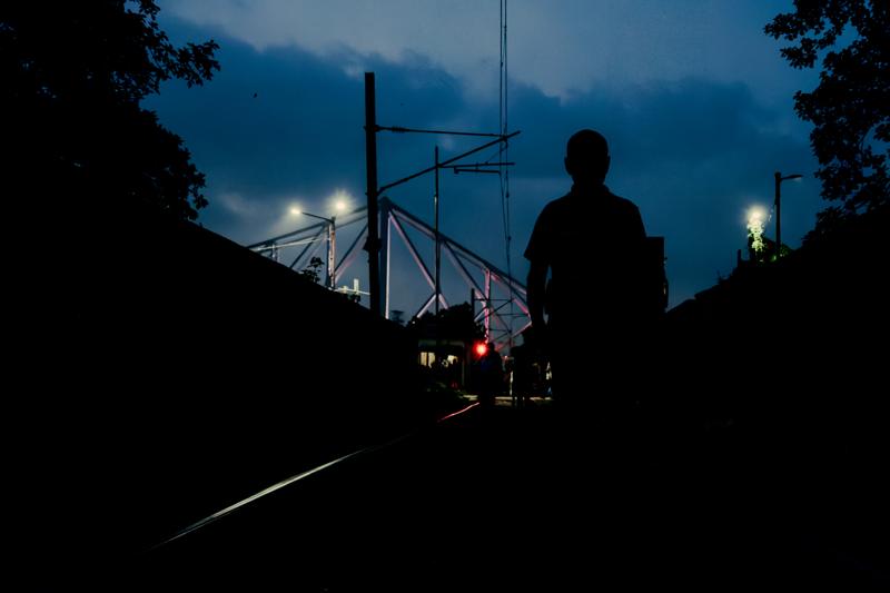 A silhouetted person stands on the road as headlights approach, with overhead wires and streetlights cutting through the blue night.