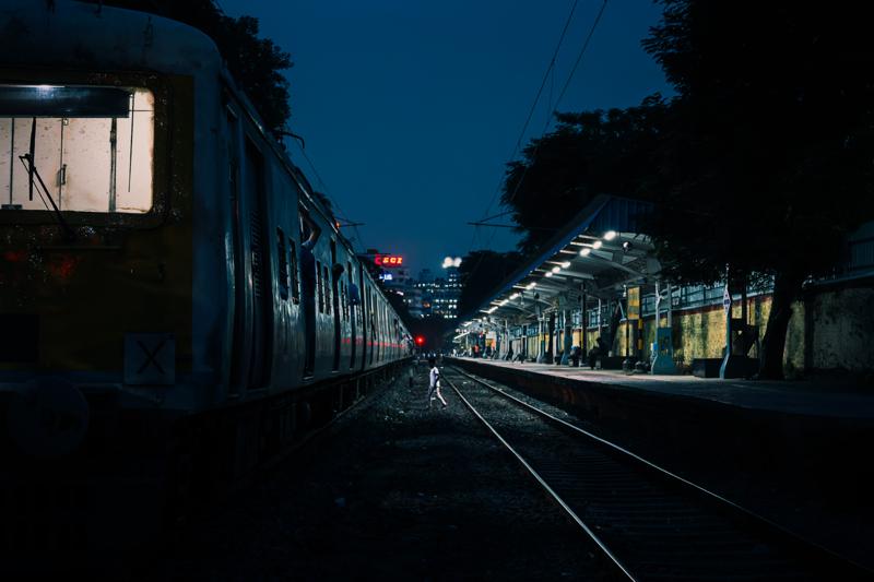 Rails converge beneath the station lamps, while a train waits at the platform and the darkness closes around the tracks.