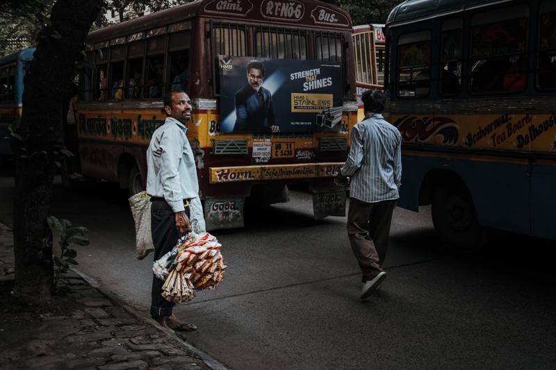Two pedestrians cross in front of a bus, one carrying a striped shopping bag, with shopfronts and traffic behind them.