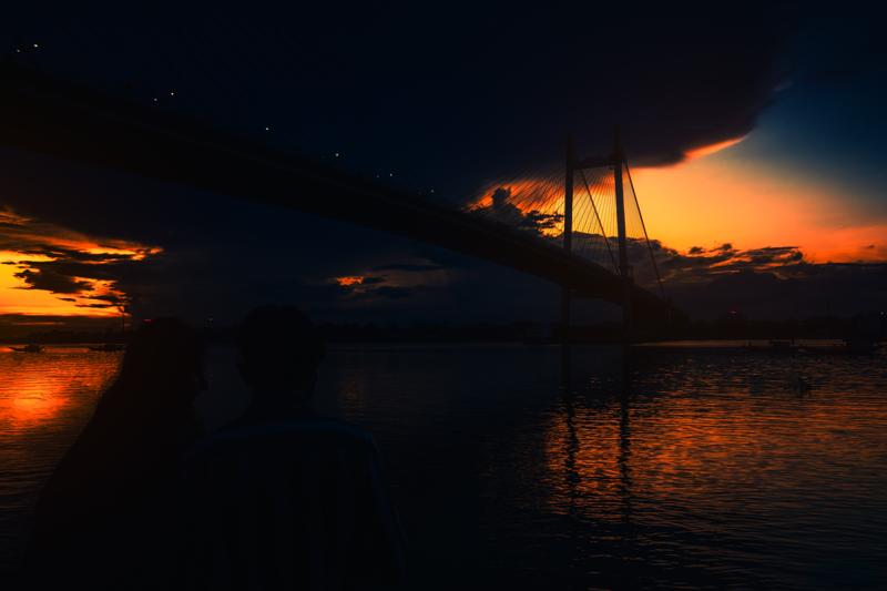 The cable bridge cuts across the blue dusk above the dark river, its warm lamps and long reflections gathering near the far bank.