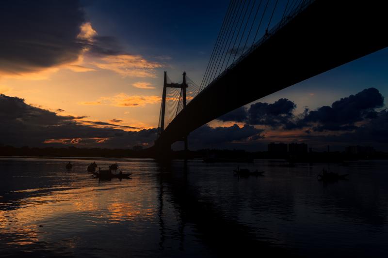 The cable bridge cuts diagonally across the blue river, its illuminated span mirrored in broken streaks below as the darkening sky gathers overhead.
