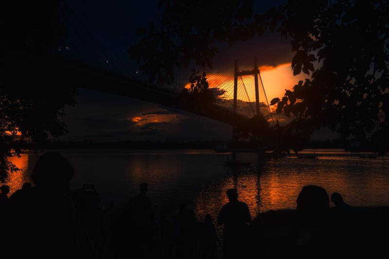 Two figures sit low beside the river as the bridge spans the glowing dusk, their dark silhouettes held against water and orange sky.