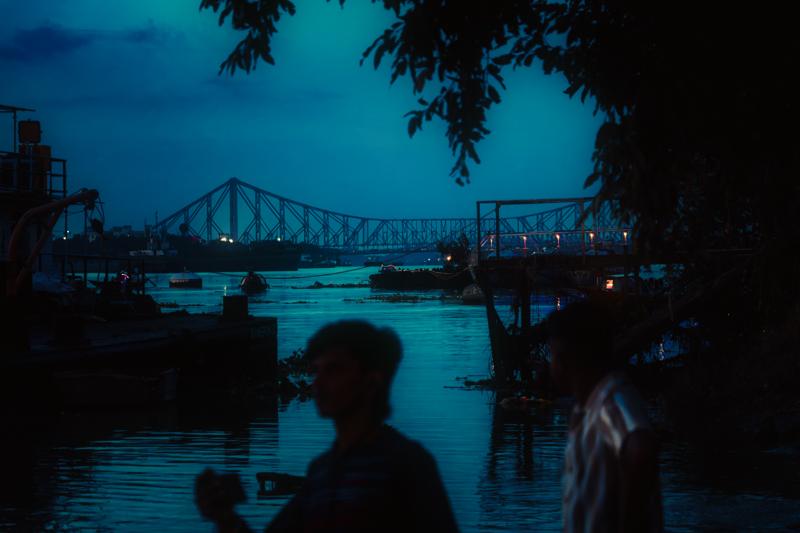 The bridge recedes into deep blue dusk above the river, a lone figure in the foreground adding scale to the wide river view.