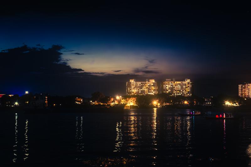 Buildings glow across the broad river beneath a deep blue sky, their broken reflections gathering in long bands on the dark water.
