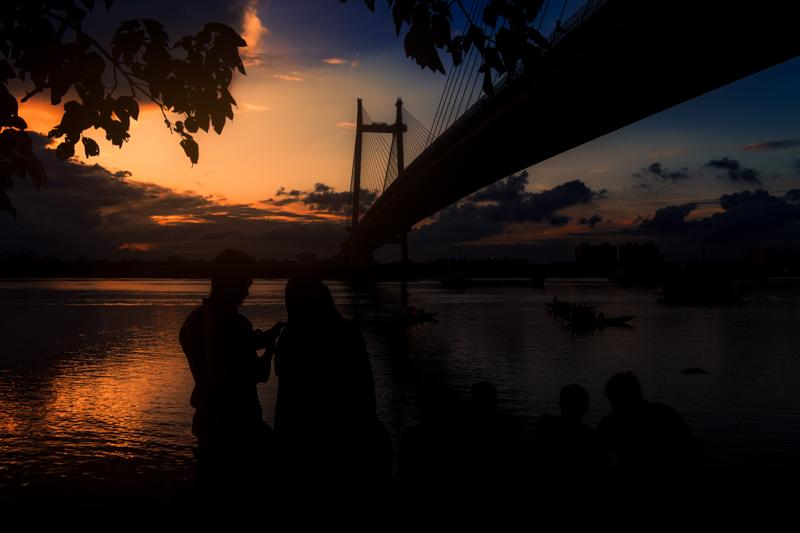A silhouetted couple faces the orange horizon from the far bank, with the steel bridge’s cable cutting across the darkening sky.
