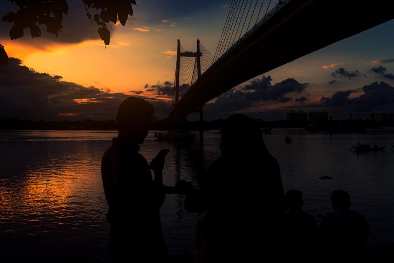 Silhouettes stand at the river’s edge beneath the steel bridge, framed by a thin orange sunset and the broader band of dark water.