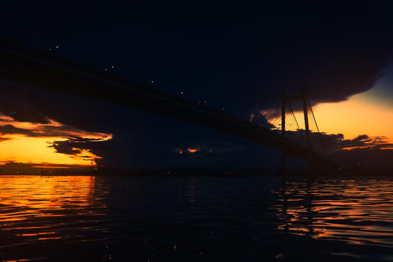 The broad river glows beneath the cable bridge as its dark deck divides the sunset, with small boats scattered along the low horizon.
