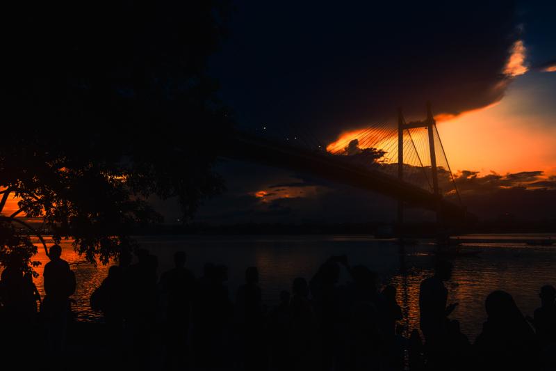 The cable bridge rises against a narrow orange sky, while its dark reflection stretches across the calm river below, nearby, from water level.