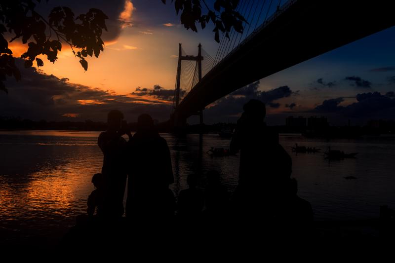 Several people stand in silhouette beneath the steel span, looking toward the broad river as the last orange light thins across the horizon.