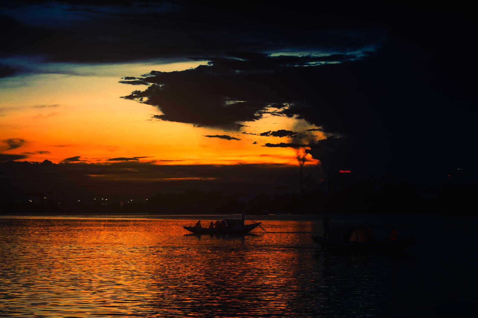 A boat rests on the Hooghly River beneath an orange sunset and deep blue clouds.