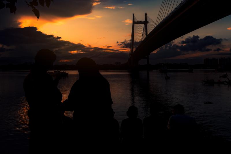A couple stands at the water’s edge as the sky burns orange behind the cable bridge, its dark span cutting across the scene.
