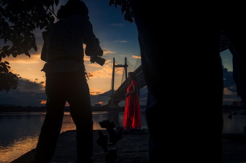Two figures stand close together beneath the steel bridge, their dark silhouettes reflected in the broad river at the end of the day.