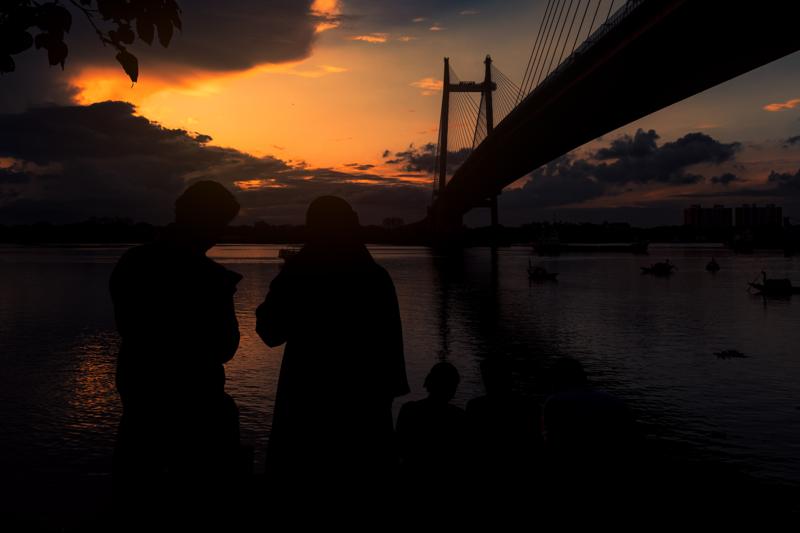 A couple stands beneath the steel bridge as sunset colours the water, the close silhouettes turning the broad view into a private pause.