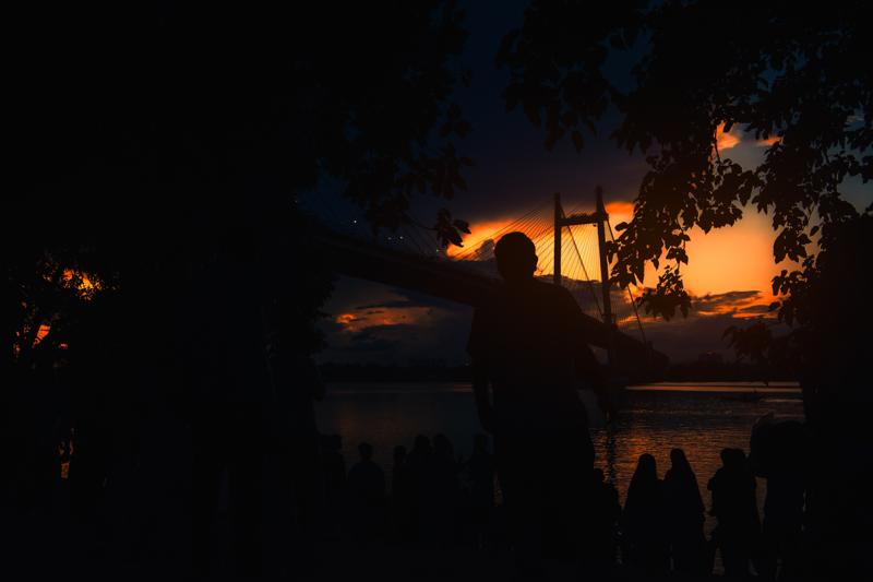 Dark figures gather near the still water below the cable span, their outlines set against the last thin strip of orange sky.