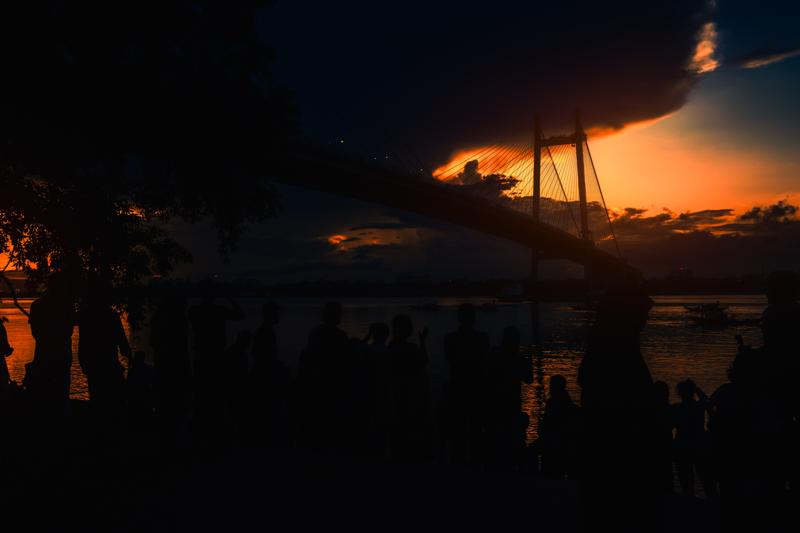 The low sun flares beside the steel bridge while a figure crosses the walkway, their silhouette briefly caught between steel and clouded sky.