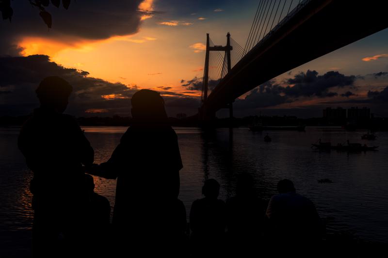A pair watches the orange sky from the far bank as the steel bridge crosses behind them, its reflection dissolving into dark water.