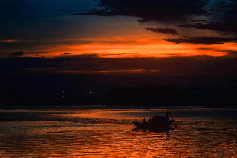 A narrow boat travels beneath a streaked orange horizon, its dark outline separating the bright band of clouded sky from the broad river.