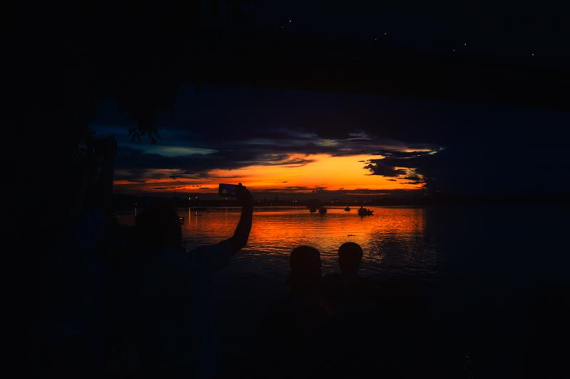 The pair stands beside the still water beneath the steel bridge, their dark outlines held against a narrow band of orange and blue.
