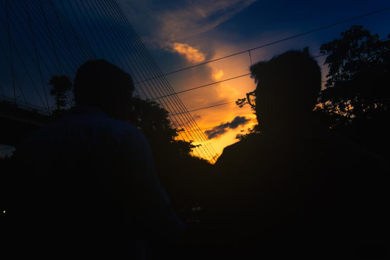Silhouettes face one another beside the still water while the cable span and its lights angle across the darkening sky, nearby, in passing.